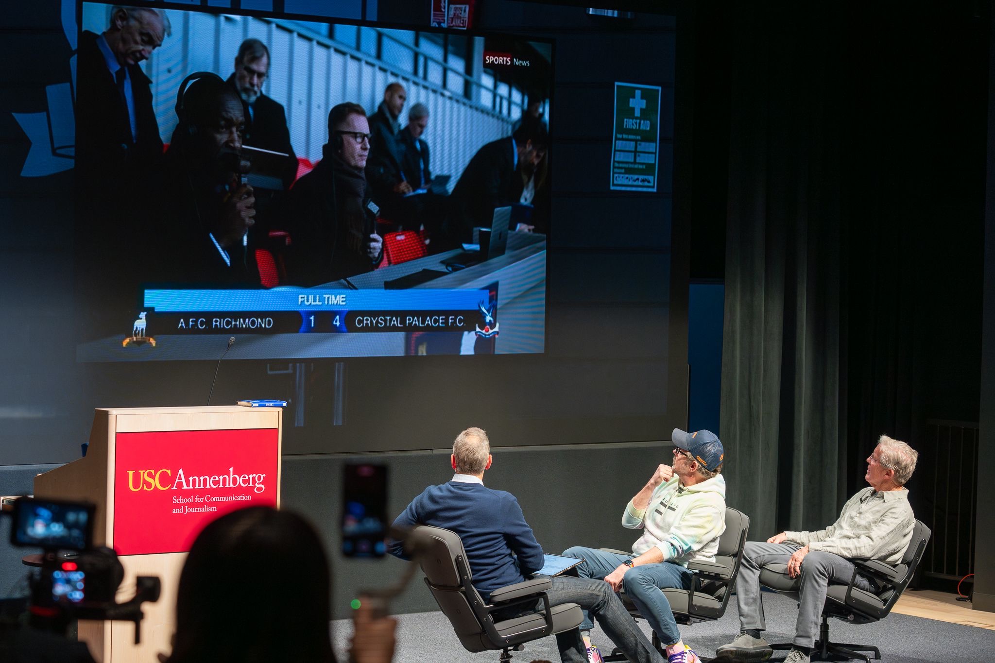 Clean three-shot of Varun Soni, Sudeikis, and Carroll relaxed and smiling on stage